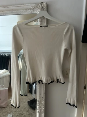 Creamvit tröja med svarta detaljer från Gina Tricot - Säljer en vit topp från Gina Tricot med långa ärmar och svarta vågiga detaljer vid ärmslut och nederkant. Tröjan har en smal passform och båtringad hals. Perfekt för dig som gillar en enkel men ändå unik stil. Använd ett fåtal gånger.