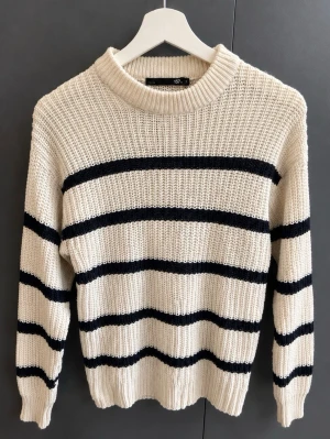 Stickad tröja med svarta ränder - Stickad tröja i vit med svarta horisontella ränder. Tröjan har en ribbad struktur, rund halsringning och långa ärmar. Perfekt för en avslappnad och trendig look. 