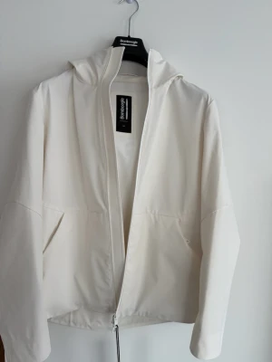 Vit vindjacka från Bomboogie - Snygg vit vindjacka från Bomboogie med minimalistisk design. Jackan har huva, dragkedja framtill och två sidofickor. Tillverkad i ett lätt material som passar perfekt för blåsiga dagar. Enkel och stilren look som funkar till det mesta.