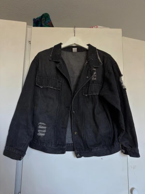 Svart oversized distressed jeansjacka - Snygg svart jeansjacka i oversized modell med slitna detaljer och råa kanter. Jackan har klassisk krage, knappstängning framtill och stora bröstfickor. Perfekt för en avslappnad streetwear-look.
