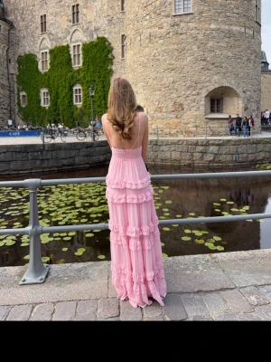 Lång rosa volangklänning - Säljer en lång, ljusrosa klänning med flera volanglager och djup v-ringning. Klänningen har smala axelband och är ärmlös, vilket ger en luftig och romantisk känsla. Perfekt för sommarens alla tillfällen. Säljer då jag hade den en gång på min bal, o nu kommer den inte till användning. ❤️