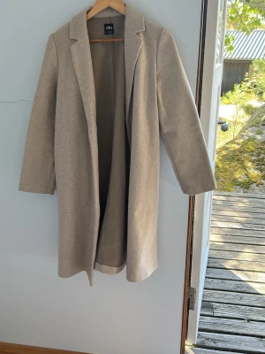 Beige kappa från ZARA - Stilren beige kappa från ZARA med klassisk krage och rak passform. Kappan är ofodrad och har en minimalistisk look utan synliga knappar eller fickor. Perfekt för dig som gillar enkel och tidlös stil.