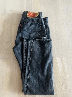 Svarta Levi's 501 jeans straight fit - Klassiska svarta Levi's 501 jeans med rak passform och hög midja. Jeansen har fem fickor, knappgylf och den ikoniska läderpatchen bak i midjan. Tillverkade i kraftig denim med vintage vibe och snygg tvätt.