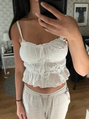 Vit topp med volang och broderi - Söt vit linnetopp med broderade detaljer, volangkant och smala axelband. Toppen har knyt framtill så man kan göra rosetter men de är egentligen ”bara” påsydda över de tre resårbanden. 