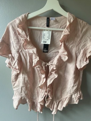Ljusrosa blus med volanger och knyt - Supersöt ljusrosa blus med volangdetaljer längs urringningen och framtill. Knytning framtill och korta ärmar ger en romantisk vibe. Perfekt till jeans eller kjol för en trendig look.