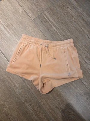 Beiga velourshorts från Juicy Couture - Säljer ett par beiga velourshorts från Juicy Couture med snörning i midjan och dragkedjefickor. Baksidan har Juicy i glittriga stenar. Perfekta för chill dagar eller när du vill ha en retro vibe.
