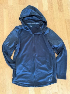 Svart hoodie från Under Armour - Snygg svart hoodie från Under Armour med dragkedja framtill och huva. Tillverkad i ett sportigt funktionsmaterial med meshfoder i huvan och glansiga detaljer på axlarna. Perfekt för träning eller chill.