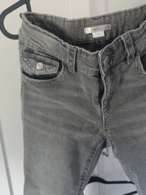 Gråa boot cut jeans med fickdetalj - Snygga grå jeans med boot cut och cool fickdetalj framtill. Jeansen har klassisk femficksdesign och silverfärgade knappar. Perfekta för en avslappnad och trendig look. Materialet är mjukt denim som ger en skön känsla hela dagen.