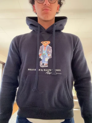 Mörkblå hoodie Polo Bear Ralph Lauren - Mörkblå hoodie från Ralph Lauren med Polo Bear-tryck på bröstet. Klassisk känguruficka, dragsko i huvan och ribbade muddar. Snygg och bekväm stil med ikoniskt björnmotiv och vit text. Perfekt för en avslappnad look.