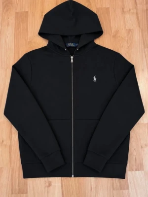 Svart hoodie från Polo Ralph Lauren - Snygg svart hoodie från Polo Ralph Lauren med dragkedja framtill och klassisk huva. Tröjan har det ikoniska Polo-logotrycket på bröstet och ribbade muddar vid ärmar och nederkant. Tillverkad i mjukt material som känns skönt mot huden.