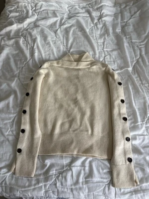 Krämvit stickad tröja med knappar - Snygg krämvit stickad tröja med svarta knappar längs båda ärmarna. Tröjan har hög krage, ribbade muddar och bred ribbad nederkant. Perfekt för dig som gillar stilrena detaljer och en mysig känsla.