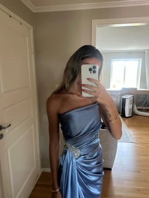 Ljusblå axelbandslös satinklänning festklänning  - Säljer en elegant ljusblå axelbandslös klänning i glansigt satintyg. Klänningen har draperad design och en snygg detalj med silverfärgade stenar i midjan. Perfekt för dig som vill sticka ut på nästa tillställning.