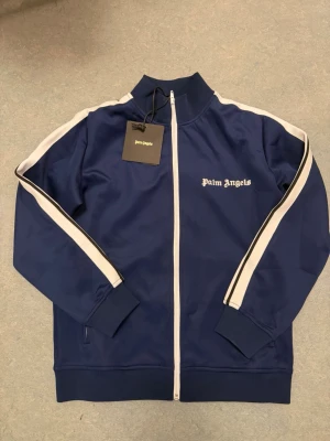 Blå ziphoodie från Palm Angels - Snygg blå track jacket från Palm Angels med vita ränder längs ärmarna och vit dragkedja. Tröjan har hög krage, ribbade muddar och logga broderad på bröstet. Tillverkad i 100% polyester för en sportig och modern look. Storlek S/M alltså passar i både S och M 