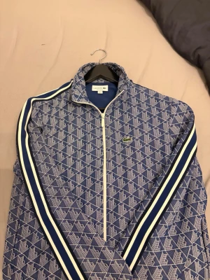 Blå mönstrad zip-jacka Lacoste - Snygg blå zip-jacka från Lacoste i regular fit med vitt och blått geometriskt mönster. Jackan har vita och marinblå ränder längs ärmarna, dragkedja framtill och klassisk Lacoste-logga på bröstet. Tillverkad i ett mjukt och bekvämt material.
