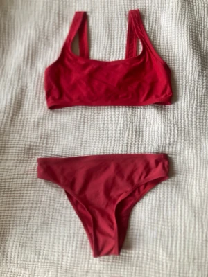 Röd bikini med sportig topp - Snygg röd bikini med enkel, sportig topp och klassiska bikinitrosor. Båda delarna är enfärgade och har en stilren design utan extra detaljer. Perfekt för lata dagar på stranden eller poolen.