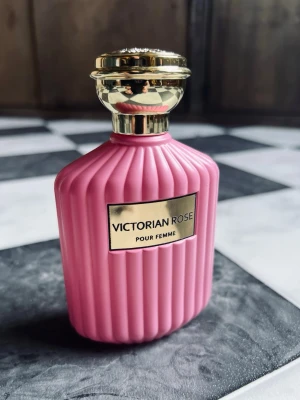 Victorian Rose Pour Femme parfym 100ml, Wadi al Khaleej - Elegant parfymflaska i matt rosa med guldfärgat lock och dekorativt mönster på toppen. Flaskans design är inspirerad av vintage och har räfflade sidor samt en metallisk etikett framtill med texten 'Victorian Rose Pour Femme'. Perfekt för dig som gillar romantiska dofter. Bara testad
