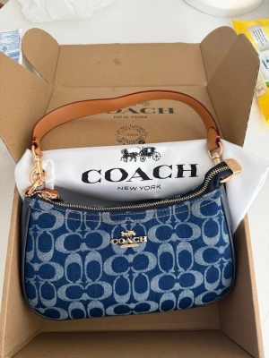 Blå monogram handväska från Coach - Snygg blå handväska från Coach med klassiskt monogrammönster i denimtyg och detaljer i brunt läder. Väskan har guldfärgade metalldetaljer och dragkedja upptill. Perfekt storlek för det viktigaste och en ikonisk accessoar till din outfit.