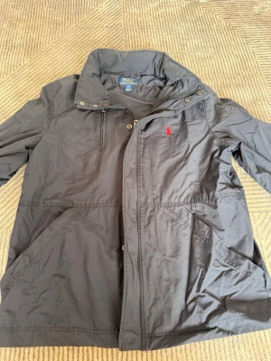 Svart vindjacka från Polo Ralph Lauren - Snygg svart vindjacka från Polo Ralph Lauren med röd broderad logga på bröstet. Jackan har hög krage, dragkedja och tryckknappar framtill samt två snedställda fickor. Tillverkad i ett lätt och slitstarkt material, perfekt för blåsiga dagar.