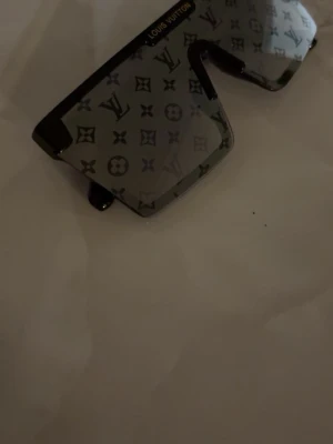 Lv solglasögon  - Säljer ett par svarta solglasögon med rektangulär sköldform och ikoniskt -mönster på glasen. Bågarna är svarta med guldfärgade detaljer vid sidorna. Perfekt accessoar för dig som vill sticka ut.