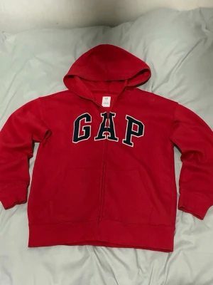 Röd hoodie med dragkedja från GAP - Snygg röd hoodie från GAP med dragkedja och stor huva. Klassisk GAP-logga i svart och vitt på bröstet. Tillverkad av minst 20% återvunnet material. Perfekt för en avslappnad och cool stil.