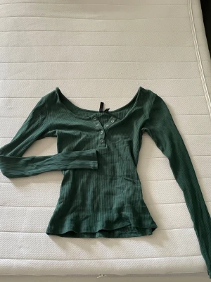 Grön ribbad topp från Pieces - Snygg grön långärmad topp från Pieces i ribbad bomullsmix som är väldigt stretchig. Toppen har en djup rundringning och knappar framtill som ger en extra detalj. 