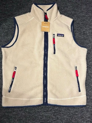 Beige fleeceväst från Patagonia - Säljer en beige fleeceväst från Patagonia med blå detaljer och röda dragkedjor. Västen har hög krage, tre fickor med dragkedja och är gjord i mjukt, fluffigt material. Perfekt för lager-på-lager och har en retrovibe med klassisk Patagonia-logga på bröstet.