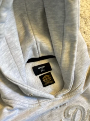 Ljusgrå hoodie från Superdry - Säljer en ljusgrå hoodie från Superdry med stor logga och text på bröstet. Hoodien har huva med dragsko, magficka och ribbade muddar vid ärmar och nederkant. Tillverkad i mjukt bomullsmaterial, perfekt för chill dagar.
