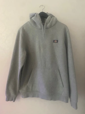 Grå hoodie från Dickies - Säljer en klassisk grå hoodie från Dickies med huva och dragsnören. Hoodien har en stor magficka och liten Dickies-logga på bröstet. Tillverkad i mjukt sweatshirtmaterial och har ribbade muddar vid ärmar och nederkant. Perfekt för en chill och avslappnad stil.