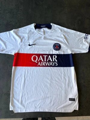 Vit PSG matchtröja från Nike - Vit PSG matchtröja från Nike med broderat klubbmärke på bröstet och Qatar Airways.  Tröjan har en bred röd och marinblå rand över bröstet och korta ärmar. Använd ett par ggr så i väldigt fint skick. 