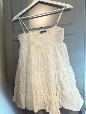 Vit kort sommarklänning med smala band - Luftig vit kort klänning med smala axelband och rynkad överdel. Klänningen har volangig kjol och är tillverkad i ett lätt material, perfekt för varma dagar. Enkel och stilren design som passar till många tillfällen. Finns en liten fläck på baksidan (se bild 3) 