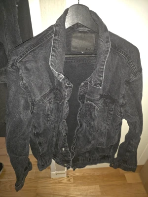 Svart oversized jeansjacka XL - Snygg svart jeansjacka i oversized modell med klassiska bröstfickor och knappstängning framtill. Jackan har en tvättad look och markerade sömmar som ger en cool vibe. Perfekt att slänga över en hoodie eller t-shirt för en avslappnad stil.