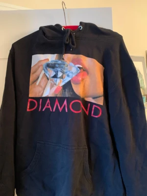 Svart hoodie med diamantmotiv - Svart oversized-fit hoodie med print på bröstet som visar en person med röda läppar och en stor diamant. Under motivet står det 'DIAMOND' i röd text. Hoodien har huva med dragsko och magficka framtill. Supply.Co! 
