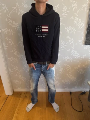 Marinblå hoodie från Lexington - Säljer en marinblå hoodie från Lexington med tryckt logga och amerikansk flagga på bröstet. Klassisk huva och ribbade muddar vid ärmslut och nederkant. Perfekt för en avslappnad och stilren look.