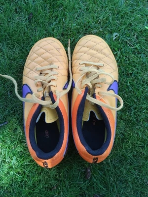 Gula Nike Tiempo fotbollsskor - Säljer ett par gula Nike Tiempo fotbollsskor med lila detaljer och svarta insidor. Skorna har klassisk snörning och ovandel i syntetmaterial med quiltad struktur för bättre bollkontroll. Perfekta för dig som vill sticka ut på planen!