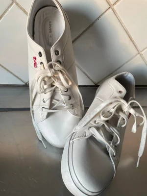 Vita låga sneakers från Levi's - Stilrena vita sneakers från Levi's med klassisk låg profil och vita snören. Skorna har diskret Levi's-logga på sidan och slitstark gummisula. Tillverkade i syntetmaterial med metallöljetter för snörningen. Perfekta för en clean och enkel look.