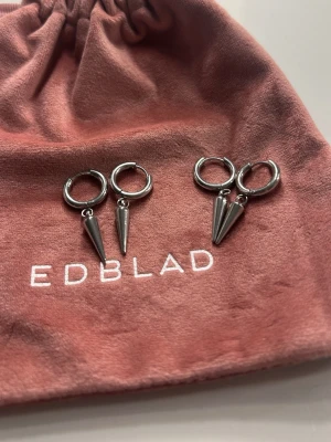 Silvriga hoops med konformad berlock - Säljer ett par silvriga örhängen från Edblad med rund hoop och en hängande konformad berlock. Örhängena har en modern och edgy look, perfekta för att lyfta din stil. Kommer i en snygg rosa tygpåse med Edblad-logga.