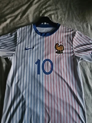 Frankrike fotbollströja Mbappé #10 - Säljer en vit fotbollströja från Nike med tunna blå och röda ränder, Frankrikes landslagsmärke och nummer 10 på bröstet. På ryggen står det Mbappé och nummer 10 i blått. 