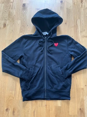 Svart cdg zip - Snygg svart zip med dragkedja och huva. På bröstet sitter det klassiska röda hjärtat som symboliserar märket cdg. Zippen har fickor framtill och justerbara snören i huvan. Perfekt för en avslappnad och trendig stil. Såklart äkta, öppen för byten och pris ej hugget i sten! 