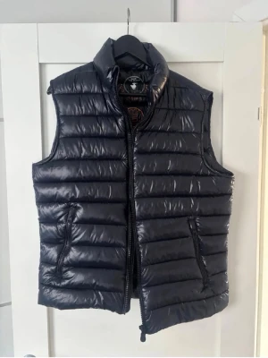 Svart dunväst från Save The Duck - Snygg svart dunväst från Save The Duck med hög krage och dragkedja framtill. Västen är quiltad och tillverkad av 100% återvunnet material, både tyg och stoppning. Två fickor med dragkedja på sidorna och diskret logga på insidan. Använd 3-5 gånger. Nyskick