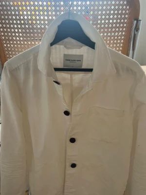 Vit overshirt från These Glory Days - Säljer en stilren vit overshirt från These Glory Days i linne. Skjortan har klassisk krage, bröstficka och mörka knappar framtill. Perfekt för en clean och avslappnad look. Liten i storleken