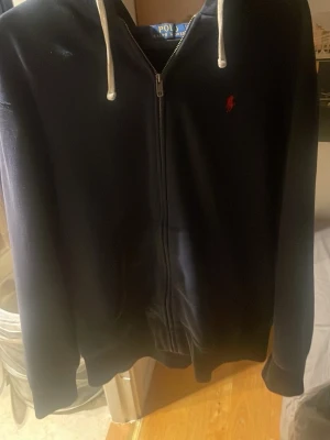 Mörkblå hoodie från Polo Ralph Lauren - Snygg mörkblå hoodie med dragkedja från Polo Ralph Lauren. Klassisk design med vit dragsko i huvan och röd broderad logga på bröstet. Mjuk bomullskänsla och kängurufickor framtill. Perfekt för en chill och stilren look.