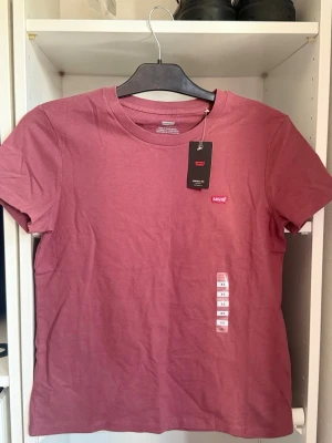 Rosa t-shirt från Levi's - Säljer en klassisk rosa t-shirt från Levi's med rund halsringning och liten broderad logga på bröstet. T-shirten har korta ärmar och är tillverkad i mjuk bomull för en skön känsla. Perfekt till jeans eller shorts.