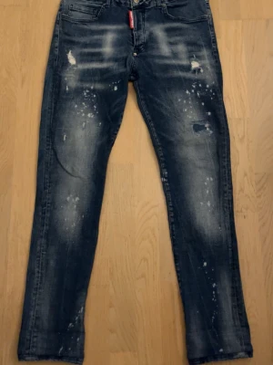 Mörkblå dsquared2 jeans  - Säljer ett par mörkblå jeans med slitningar och färgstänk för en cool, trendig look. Jeansen har raka ben, klassiska fem fickor och ett färgglatt patch med texten 'ICON' bak i midjan. Perfekta för dig som gillar streetwear och vill sticka ut.