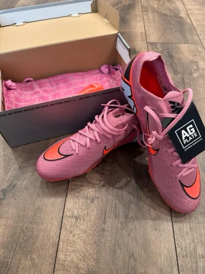 Rosa Nike fotbollsskor AG Plate - Snygga rosa Nike fotbollsskor med AG Plate för spel på konstgräs. Skorna har en strömlinjeformad design, svart och orange Nike-logga samt snörning för bra passform. Yttermaterialet är syntetiskt och sulan har dobbar anpassade för konstgräs.