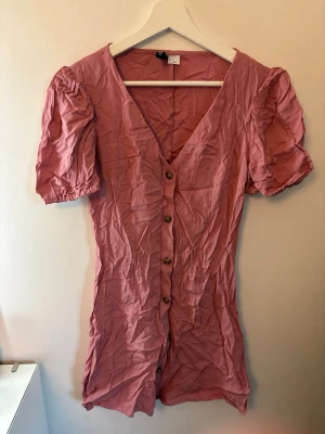 Rosa klänning med puffärm och knappar - Kort rosa klänning med puffärmar och v-ringning. Klänningen har knappar framtill och är gjord i ett lätt material, perfekt för varmare dagar. Puffärmarna har resår vid ärmslutet för en söt look.