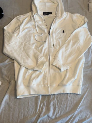 Vit hoodie från Polo Ralph Lauren - Säljer en vit hoodie från Polo Ralph Lauren med dragkedja framtill och klassisk huva med snören. Tröjan har det ikoniska mörkblå logobroderiet på bröstet och ribbade muddar vid ärmslut och nederkant. Perfekt för en clean och stilren look.