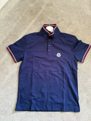 Marinblå Moncler pikétröja - Snygg marinblå pikétröja från Moncler med klassisk krage och knäppning framtill. Tröjan har röda och vita ränder på krage och ärmslut samt Moncler-logga på bröstet. Perfekt för en stilren och sportig look.