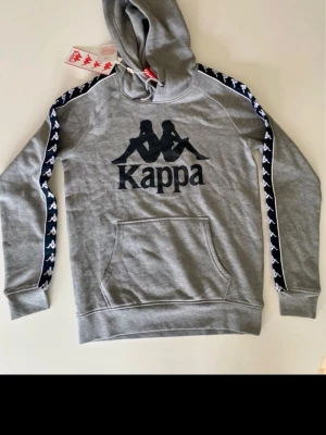 Grå hoodie från Kappa med logga - Grå hoodie med svart Kappa-logga på bröstet och svarta/vita band längs ärmarna. Hoodien har magficka, huva med dragsko och är tillverkad i mjukt sweatshirtmaterial. Perfekt för en sportig och avslappnad stil.