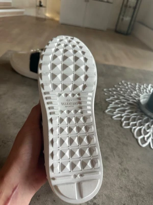 Vita sneakers från Valentino - Snygga vita sneakers från Valentino med svart detalj över sidan och klassisk snörning. Skorna har en låg profil och en mönstrad sula i vitt gummi. Perfekta för dig som vill ha en stilren och trendig look. Säljs pga det var för små 
