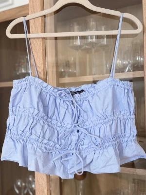 Ljusblå croppad topp med snörning - Söt ljusblå croppad linnetopp med tunna axelband och snörning framtill. Toppen har volangkant nedtill och rynkade detaljer.